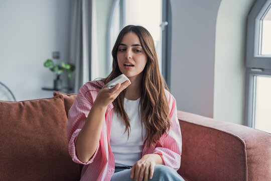 A happy young woman relaxes on a comfortable sofa at home, smiling as she records and sends an audio message to friends, enjoying a peaceful weekend while using her voice assistant