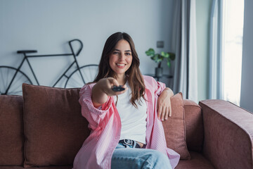 Naklejka premium A cheerful young woman lounges on a living room sofa, delighted by a TV program, embracing leisure time, comfort, and the relaxed atmosphere of a cozy home weekend