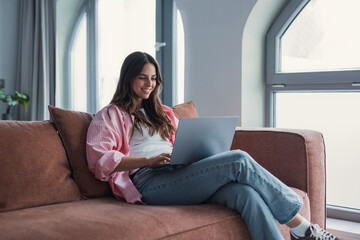 A focused young millennial woman sits on a sofa at home, working on her laptop and completing...