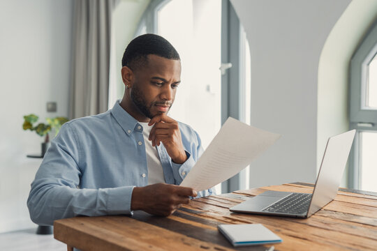Concentrated African American manager reading letter, paper handout, analyzing company statistics, thinking about financial risks, comparing results. Thoughtful black worker considering business offer