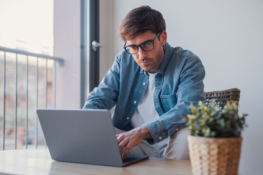 Side view handsome young businessman working with computer remotely, sitting at wooden table in office. Pleasant happy man communicating in social network, searching information online.