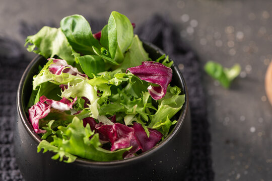 A vibrant blend of fresh green fris&eacute;e and purple radicchio leaves served in a sleek black bowl the salad sits on a dark textured napkin with a soft bokeh background showing salt and pepper