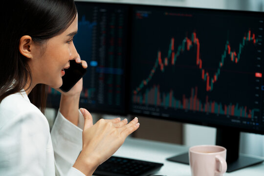 Young beautiful Asian businesswoman calling to broker on phone to analyze stable or higher profit value with profitable each exchange stock market graph on pc screens at modern office. Stratagem.
