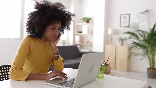 Smiling young woman with afro hair sitting at her desk in a modern living room, working remotely, browsing social media, or e-learning with her portable computer in a bright interior