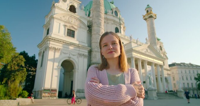 Cinematic hero shot of a confident young Caucasian woman standing with arms crossed in front of the Karlskirche in Vienna, Austria. Proud female traveler posing before the Baroque cathedral