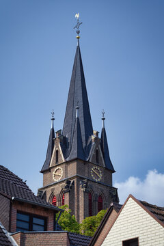 Scenic view of the tall brick spire of St. Laurentius in Ensen, Cologne, featuring golden clocks and a weathercock against a clear blue sky