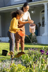 Diverse mother and daughter watering flowerbed at front yard with pink and orange watering cans