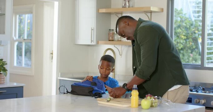African American dad, son packing school lunch at kitchen island, father placing box into backpack