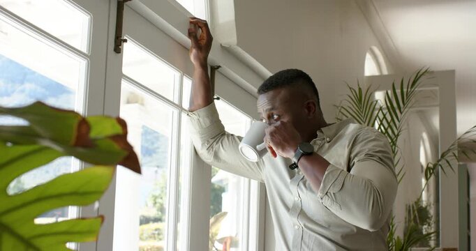 Sunlight is pouring, left arm resting, hand sipping white mug while peering at hills through window