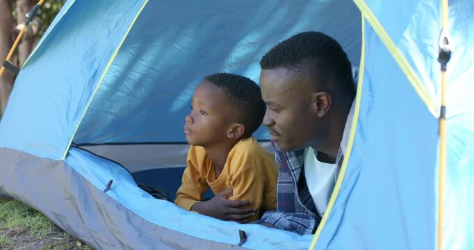 African American dad and son hearing rustle, lying at blue tent entrance watching, boy in yellow