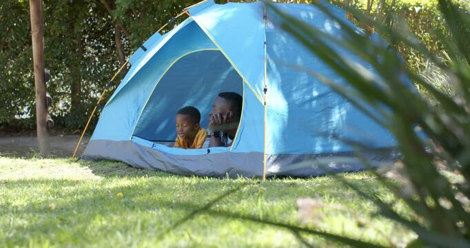 African American father-son sitting in tent on lawn, father holding smartphone taking incoming call