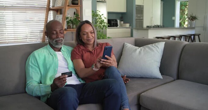 Senior couple on gray-sofa man pointing credit card leading to woman tapping blue smartphone to pay