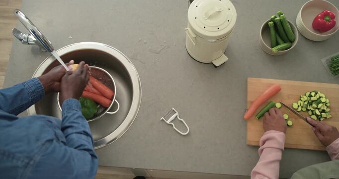 Diverse couple starting meal prep rinsing carrots in colander and slicing zucchini at home counter