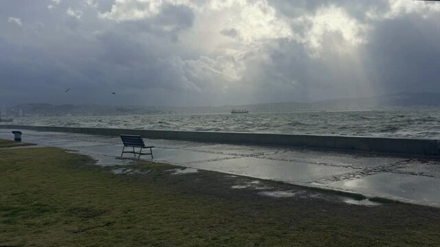 Powerful sea waves crash against a coastal promenade under dramatic storm clouds. Empty bench, flying seagulls and rough water create a moody seaside atmosphere before rain.