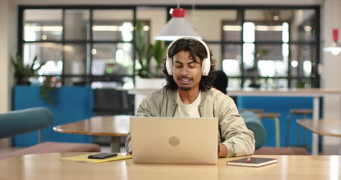 Indian man hearing audio with headphones, joining meeting while typing on laptop at communal table