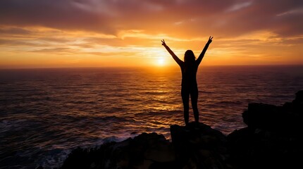 Fototapeta premium Woman silhouette with raised arms celebrating success at ocean sunset from rocky cliff with golden sky and waves below for motivation concepts.