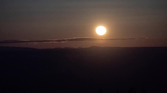 Full moon rising through a thin cloud over a distant mountain ridge at dusk, time lapse