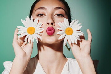 Naklejka premium Young woman holding daisies near face with bright lipstick posing on green background. Concept of beauty, fashion, creative, fresh, and playful portrait with natural flowers.