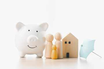 Piggy bank with family figures, wooden house and small umbrella on bright desk, symbolizing savings, home insurance, financial planning, mortgage security, risk protection and future stability.