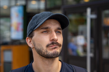 Close up portrait of a young bearded man wearing a black baseball cap and small hoop earring, gazing upward with a calm thoughtful expression, in front of a softly blurred city entrance and street  © Masarik