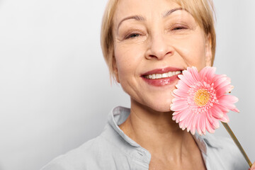 Smiling mature woman holds pink gerbera flower near face, wearing light shirt on white background....