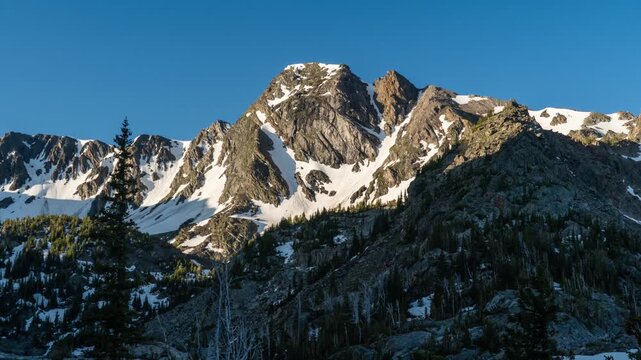 Morning sunlight sweeping down a rugged snowy mountain peak above a dark forest, Gallatin Range, Montana, time lapse