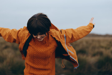 Happy woman lifestyle outdoors wearing orange sweater and jacket spreads arms wide in nature field....