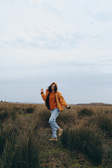 Fototapeta premium Young woman lifestyle walking outdoors in casual autumn clothing with coffee cup in hand. Natural environment with grass and cloudy sky creates relaxed and cozy mood for daily routine.