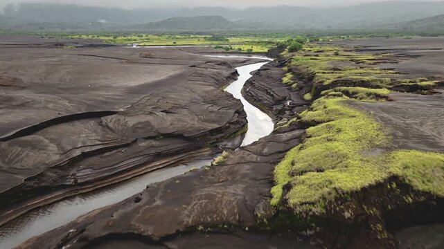 Volcanic ash field land defining rugged, barren landscape on Tanna Island, winding river cutting deep gorge through dark, layered terrain towards the distant lush green vegetation. Aerial drone flight