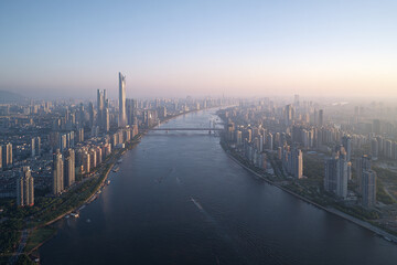 Fototapeta premium Aerial view of modern city skyline with high rise buildings along wide river, bridges crossing water, and hazy sky at sunset creating calm atmosphere