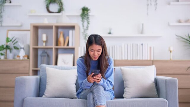Happy young asian woman relax on comfortable couch at home, typing chat message on smartphone, smiling girl use cell phone chatting, searching information on browser wireless internet, online shopping