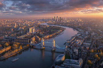 Fototapeta premium Cityscape panorama of London with River Thames and Tower Bridge at sunset, showing urban buildings, skyscrapers, and dramatic cloudy sky creating warm, peaceful atmosphere