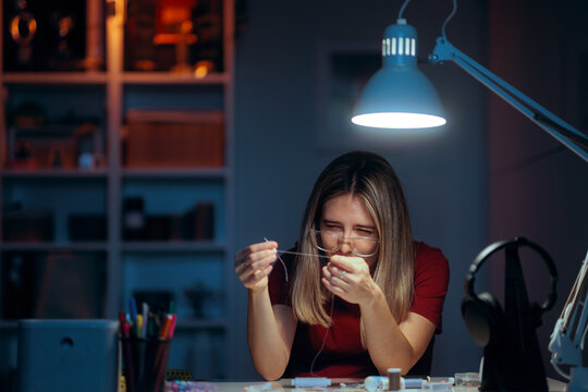Woman with Eyeglasses Struggles to Thread a Needle under Lamp Light. Seamstress wearing glasses having difficulty threading a needle in low light 