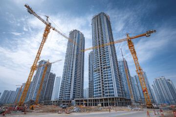Naklejka premium Construction site with multiple high rise apartment buildings under construction and several large yellow cranes against cloudy blue sky, showing urban development progress