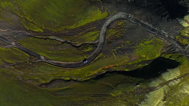 Top-down drone view of a tiny car on a winding highland road above a dark volcanic lake, surrounded by green moss and rugged terrain. Remote summer travel and Nordic adventure.