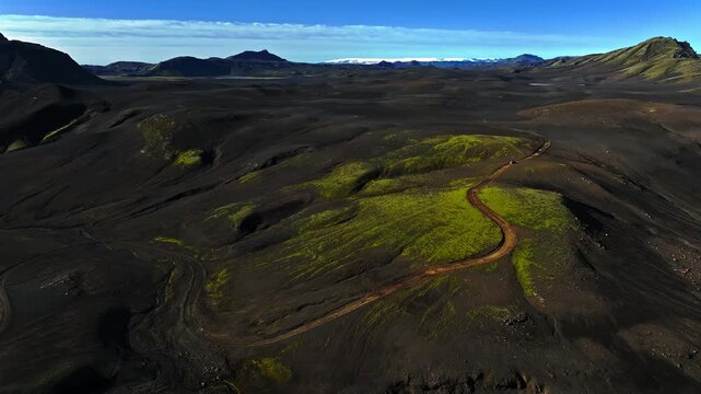 Drone panorama of a winding dirt road across black volcanic hills with bright green moss patches in Iceland. A joyful long journey vibe through remote summer wilderness.
