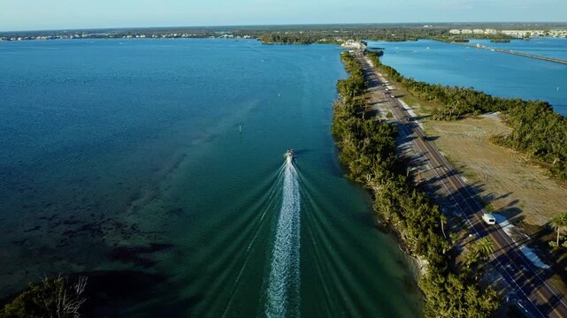 A small boat moves through a clear coastal waterway alongside a tree-lined shore and a parallel highway, with distant residential buildings visible under a clear sky in Clearwater, Florida.