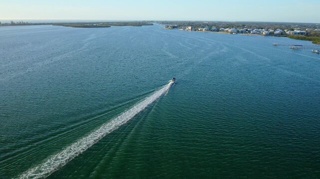 A speedboat travels through a wide coastal channel lined with homes and docks, leaving a white water trail behind as it moves steadily across calm water on a clear day.