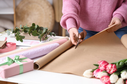 Young woman with gift box and packing materials cutting wrapping paper for tulip flowers bouquet at white table. International Women's Day