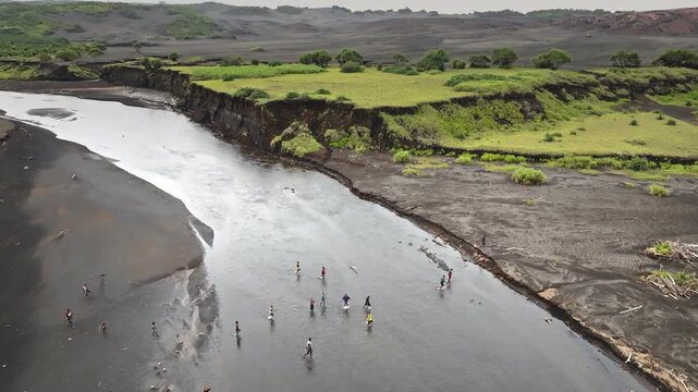 People walking along shallow river flowing through black volcanic sand landscapes, surrounded by green coastal vegetation and eroded riverbanks in Port Resolution, Tanna Island, Vanuatu. Aerial view
