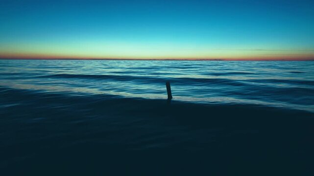 Floating object partially submerged in dark ocean waters as orange hues of sunset glow on the horizon, with waves breaking softly during evening twilight.