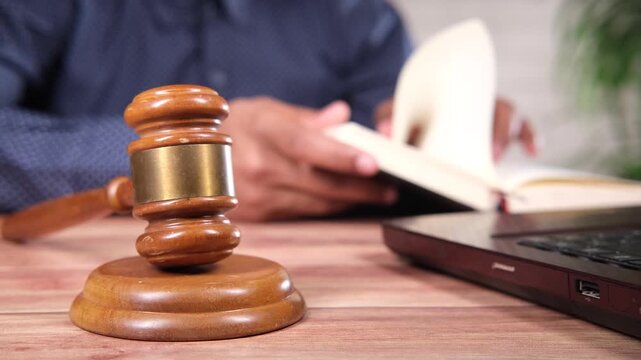Gavel resting on wooden table while person in background studies law book next to laptop, symbolizing justice and legal education