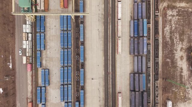 Top down aerial view of a large industrial freight terminal with rows of blue shipping containers and parallel railway tracks.