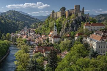 Obraz premium Medieval stone castle on rocky hill overlooking peaceful river and charming European town with red rooftops and lush green forest under blue sky with clouds
