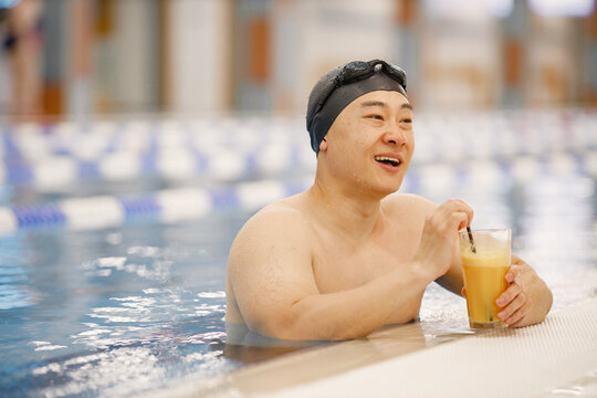 Asian man in hat and goggles drinking an orange juice in swimming pool