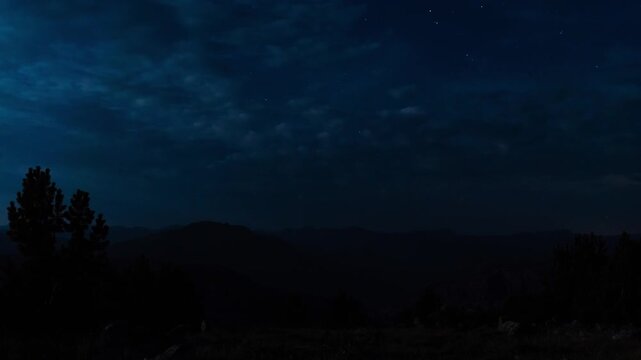 Stars and clouds over the dark silhouette of the Absaroka Range and pine trees at night, Montana, time lapse
