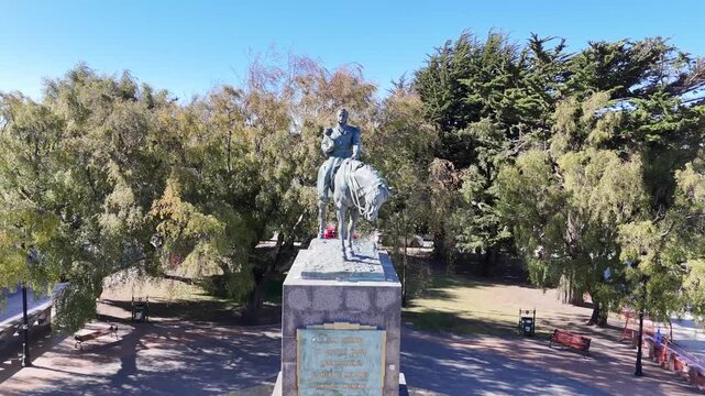 Aerial pulls back from the bronze equestrian statue of General Manuel Bulnes to reveal the entire surrounding park, featuring empty benches, paved pathways, and lush green cypress trees