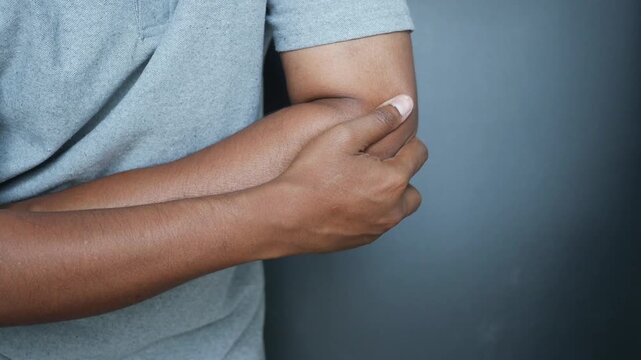 A close up view of a man in a gray t-shirt experiencing elbow pain, showing discomfort and holding the affected area