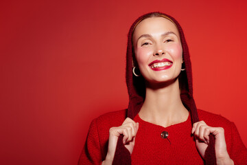 Smiling woman in red sweater and hood poses against red background. She shows happiness and...