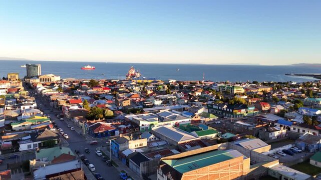 Aerial lies forward over the colorful houses and industrial buildings of Punta Arenas, Chile, moving toward the coast where ships are docked in the blue waters of the Strait of Magellan